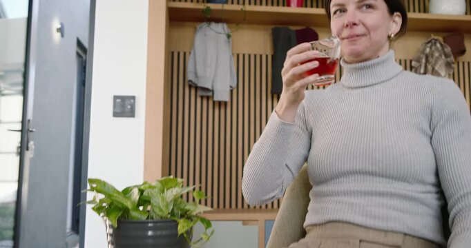 Woman seeing phone message by door, smiling, reaching for glass, sipping, looking around to relax