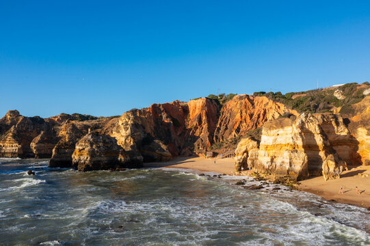 Golden sea cliffs and a sandy beach line the Algarve coastline near Lagos as waves break along the shore under a clear blue sky.