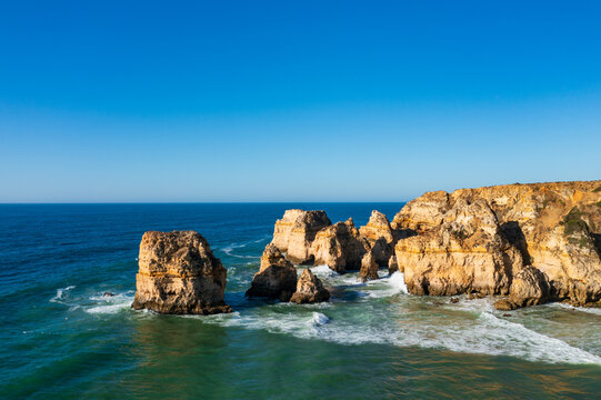 Rocky sea stacks and golden cliffs rise above the Atlantic Ocean along the Algarve coast as waves break under a clear blue sky.