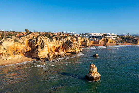 Golden cliffs and a sandy beach line the coast, with a rocky shoreline, clear blue water and low-rise buildings in the distance.