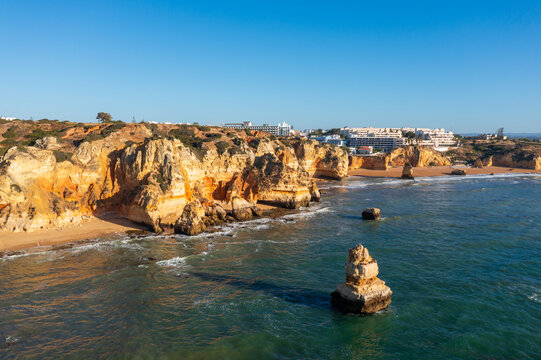 Rocky sea cliffs and a sandy beach line the coast of Lagos, with a calm ocean and a clear blue sky.
