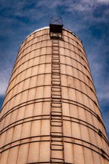 Tall vintage concrete grain elevator silo with metal ladder climbing to the top against a deep blue sky with light clouds in low angle view © Donald