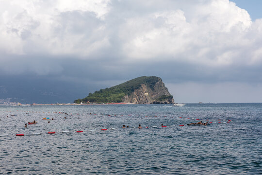 Tourists swim in the Adriatic Sea near the rocky Sveti Nikola island along the coast of Budva municipality in Montenegro under a cloudy sky during summer season