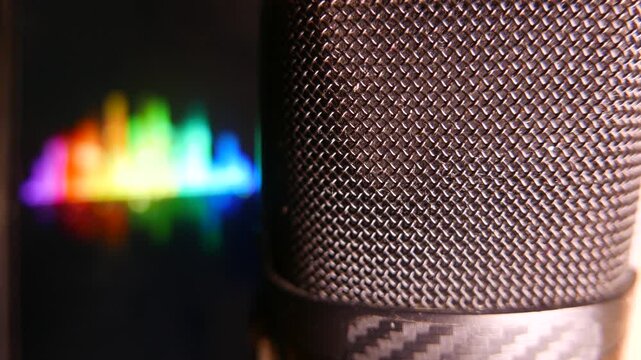 Close-up of a professional microphone in an audio recording studio. Colorful sound levels move on a computer screen in the background. POV: recording audio; recording a podcast; recording a voiceover.
