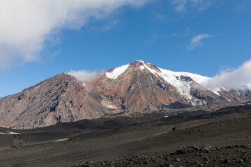 Scenic view of the Plosky Tolbachik volcanic mountain with snowy slopes and volcanic ash plateau in Kamchatka Krai, Russia on a sunny summer day under bright blue sky with clouds © TravelFlow