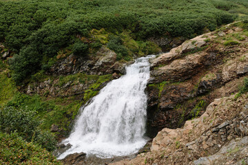 Picturesque mountain waterfall cascades over rocky cliffs surrounded by lush green slopes in the Vachkazhets mountain area of Kamchatka Krai, Russia, showing natural wilderness scenery © TravelFlow