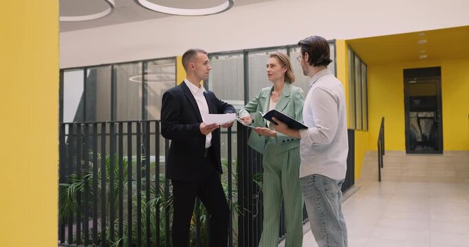 Team of three diverse business professionals colleagues discussing project on standing meeting in office atrium engaged in strategizing sharing ideas coordinating tasks using paper and digital tools