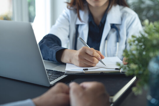 Doctor consulting patient, filling medical form on clipboard beside laptop in clinic office.