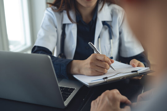 Doctor consulting patient, filling medical form on clipboard beside laptop in clinic office.