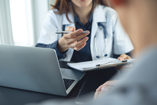 Female doctor explaining medical results to patient, holding pen over clipboard beside laptop in clinic office.