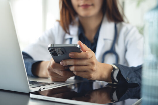 Female doctor using smartphone while working at desk with laptop and digital tablet in modern medical office.