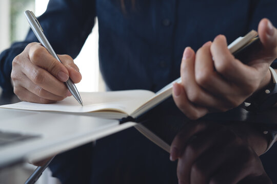 Close up, woman hand with a pen writing in notebook with laptop on office desk, planning and making a note reminder