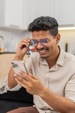 Indian man at home checks messenger long small text on smartphone puts on glasses and reads clearly. Arabian guy on sofa improves focus smiles and continues chat and scrolls through message thread