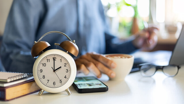 close up shot, focus alarm clock 14.00 PM with blurred businessman holding coffee cup working and meeting online with laptop on desk