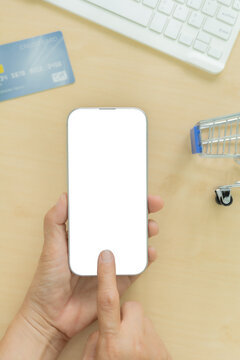 Top view of hands holding smartphone with blank white screen mockup, blue credit card, miniature shopping cart, and keyboard on wooden desk. Concept for online shopping, ecommerce, and mobile payment.