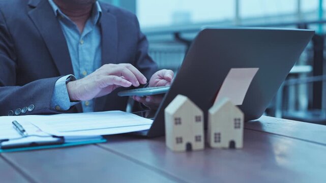 Businessman calculating mortgage loan rates beside wooden house model miniatures on outdoor desk.