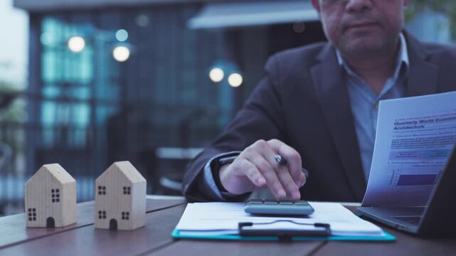 Mature businessman calculating property investment costs outdoor beside wooden house model, financial reports, laptop, pen, clipboard.