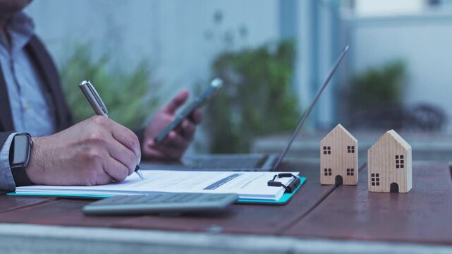 Businessman signing loan document contract beside laptop calculator miniature wooden house model outdoors desk.
