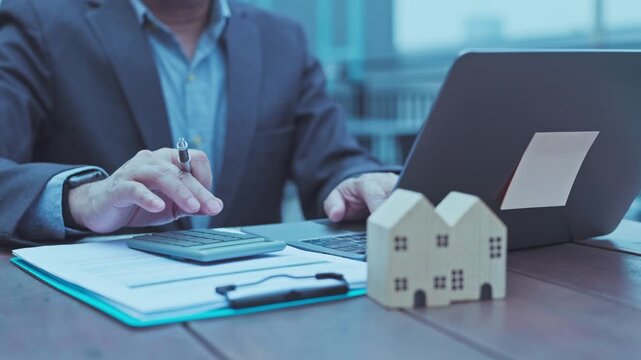 Real estate agent calculates mortgage loan rates beside wooden house model, laptop, clipboard contract papers on office desk.