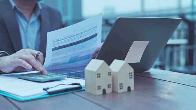 Businessman analyzing financial report document beside calculator wooden house model laptop desk calculating mortgage investment loan budget office.