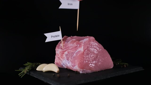 A male hand inserts protein, iron, vitamin B12 and zinc markers into pork on black slate with rosemary, garlic and pepper nearby. Meat nutrition concept