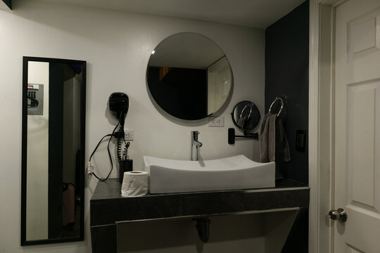 Modern bathroom vanity featuring a unique rectangular basin, chrome faucet, wall-mounted circular mirror, hair dryer, and a smaller magnifying mirror, presenting contemporary home design and hygiene