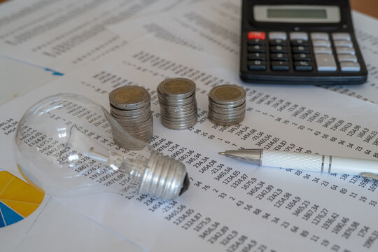 Calculating electricity consumption bills, showing a light bulb, stacked coins representing increasing costs, financial documents, a pen, and a calculator on a desk