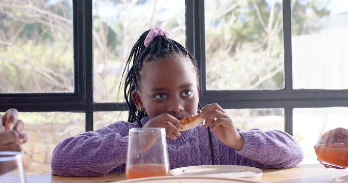 At mealtime African child nibbling pizza slice, pausing and glancing at table, purple sweater