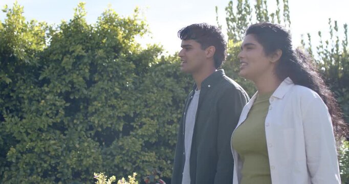 Diverse couple walking, chatting along garden path past rose bushes, tall hedge, sunlit, copy space