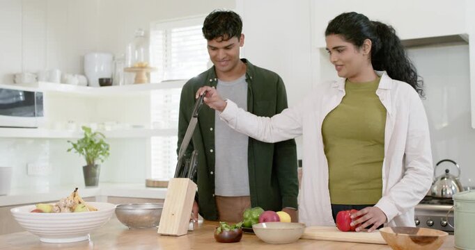 Diverse couple in kitchen, woman reaching knife block, slicing red pepper on cutting board, cooking