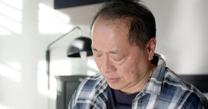 Senior Asian man wearing plaid looking toward blinds in bedroom, sunlight drawing eye to lamp
