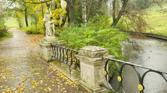 A centaur statue stands quietly beside a wet bridge in gentle rain. Camera glides in slow motion through Pavlovsk Park, Russia. The scene evokes a calm, nostalgic feeling.