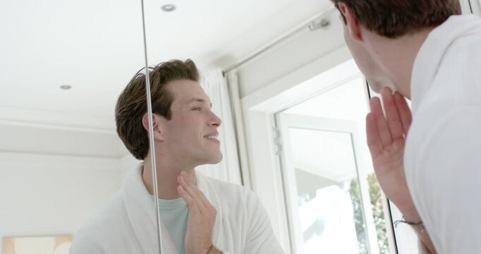 Man facing mirror checking neck, probing teeth, smoothing skin in bathroom for grooming in bathrobe