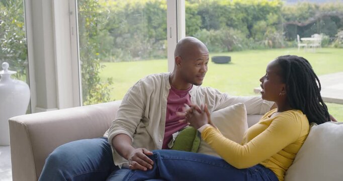 African American couple sitting on light sofa by glass doors, sharing playful talk after tease