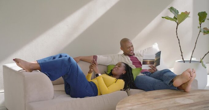 Couple on sofa, woman scrolling smartphone causing man holding booklet leaning and smiling