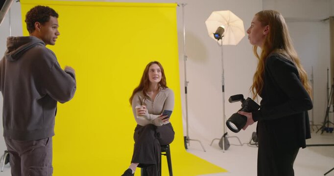 Diverse crew photog cueing shot as asst tweaking lights and model holding coffee by yellow backdrop