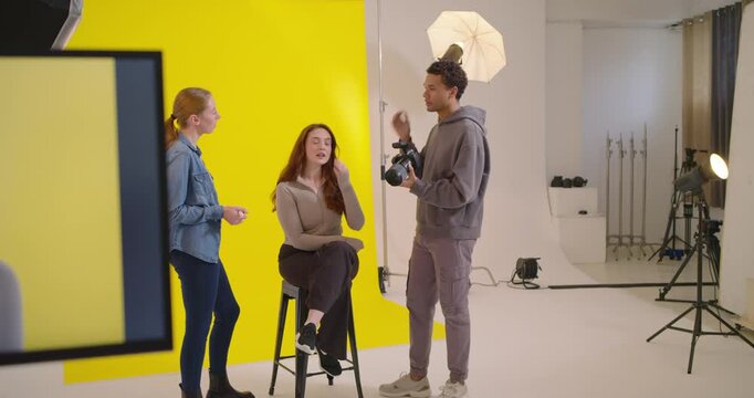 Monitor showing, stylist touching up makeup hair on female stool for portrait, photog-checking DSLR