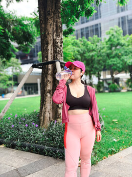 Asian woman in pink activewear drinking water from a bottle while resting after a workout in a modern urban park