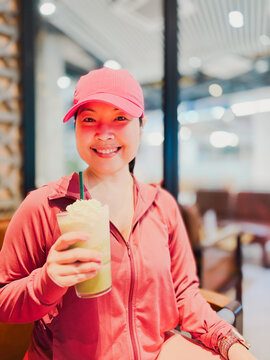 Portrait of a smiling Asian woman in pink activewear sitting in a cafe holding a cold blended matcha green tea frappe after a workout