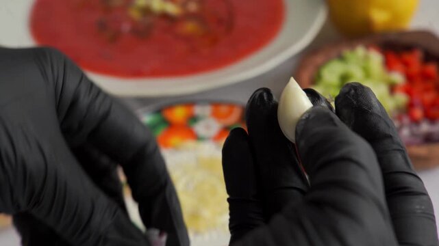 Hands in black gloves peeling a fresh garlic clove over a table with gazpacho ingredients, cooking preparation close-up