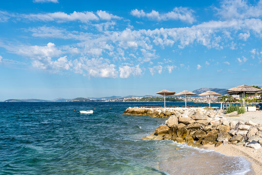 A scenic Adriatic shoreline in Croatia featuring rocky breakwaters, beach sunshades, clear turquoise water and distant coastal settlements beneath a bright blue sky filled with white clouds.