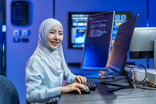 Friendly smiling young Muslim female software developer wearing a hijab, actively coding software applications on dual computer monitors inside a bright modern high-tech office environment.