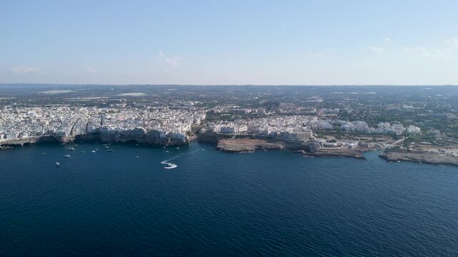 Approaching the coastal town of Polignano a Mare from the open sea, dark blue water filling the foreground and white buildings lining low cliffs on the horizon, a few boats near shore, concept of arr