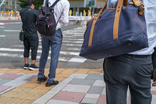 Close-up of a commuter carrying a blue canvas bag with tan leather handles over the shoulder while waiting at an urban crosswalk with pedestrians in the background.
