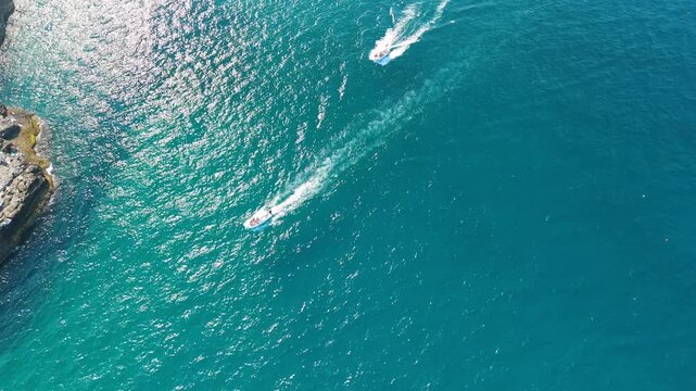 Two motorboats rounding a rocky limestone point near Polignano a Mare, their white wakes curving across sparkling deep blue water with sun glare reflecting off the waves, aerial view of busy summer s