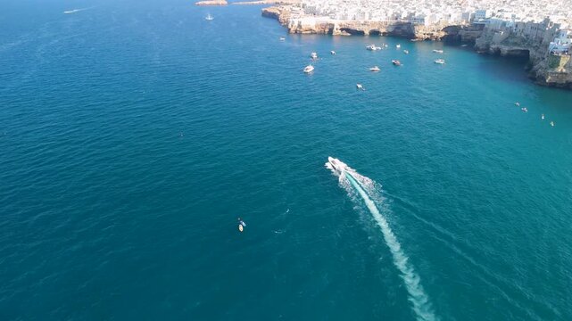 High aerial view of a speedboat heading toward a fleet of anchored boats beneath the cliffs of Polignano a Mare, its curved white wake tracing across deep blue sea with swimmers and paddle boards nea