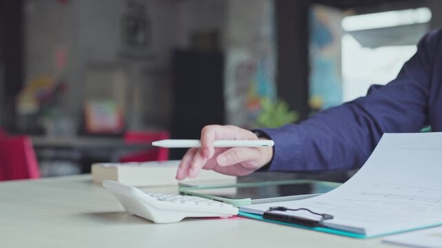 Business accountant writes notes on digital tablet screen near calculator clipboard documents inside bright workplace.
