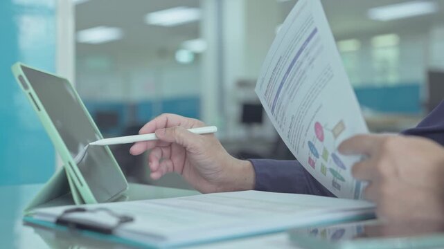 Professional analyst reviews printed system architecture diagrams operates digital tablet with pen tool in bright corporate office workspace.