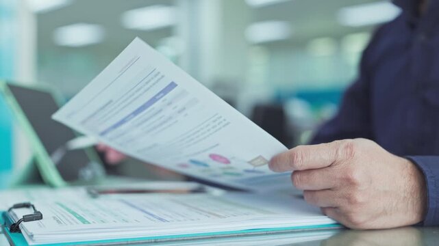 Businessman reviews printed financial document charts alongside tablet computer on desk in corporate office space.