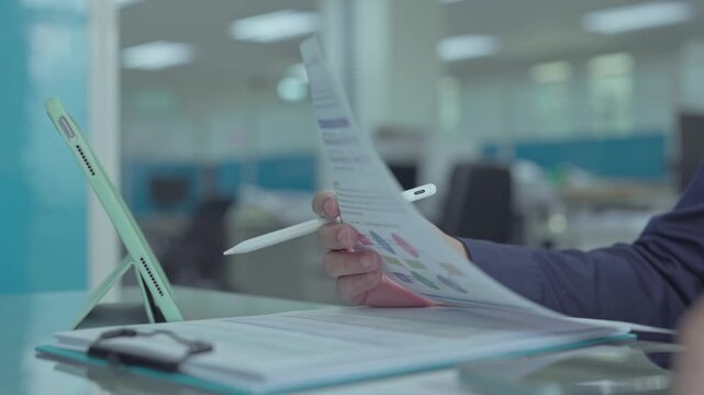 Auditor holding financial documents digital pen reviews printed report sheets near tablet computer inside corporate office workplace.